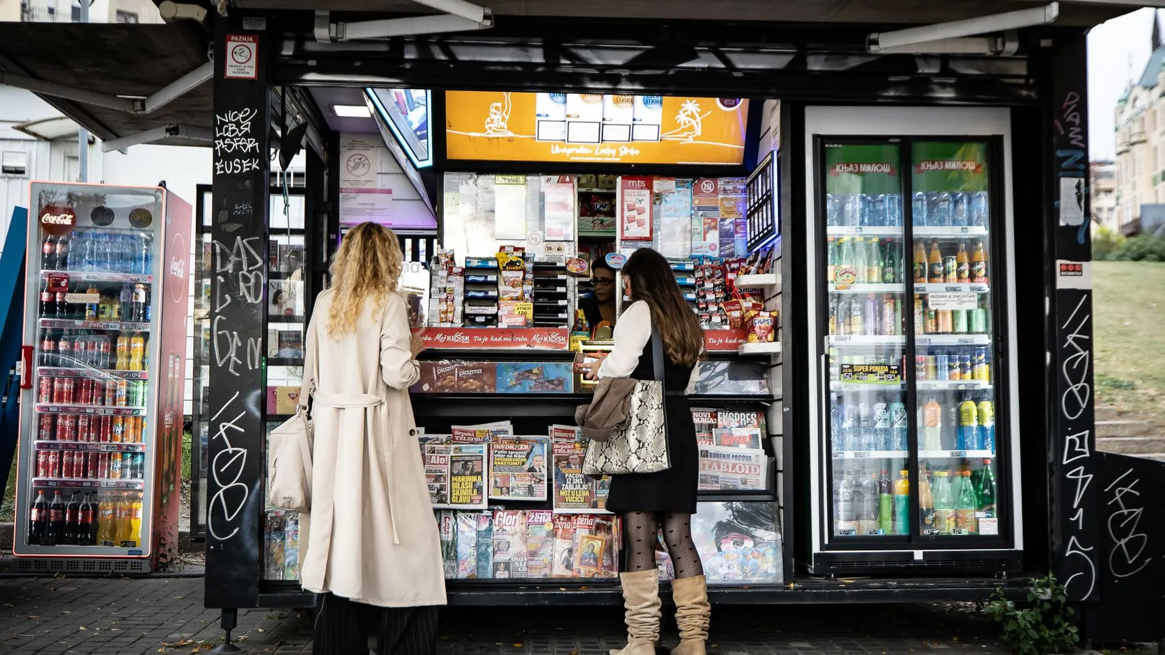 Deux femmes devant un kiosque de rue garni de journaux, de friandises et de réfrigérateurs à boissons