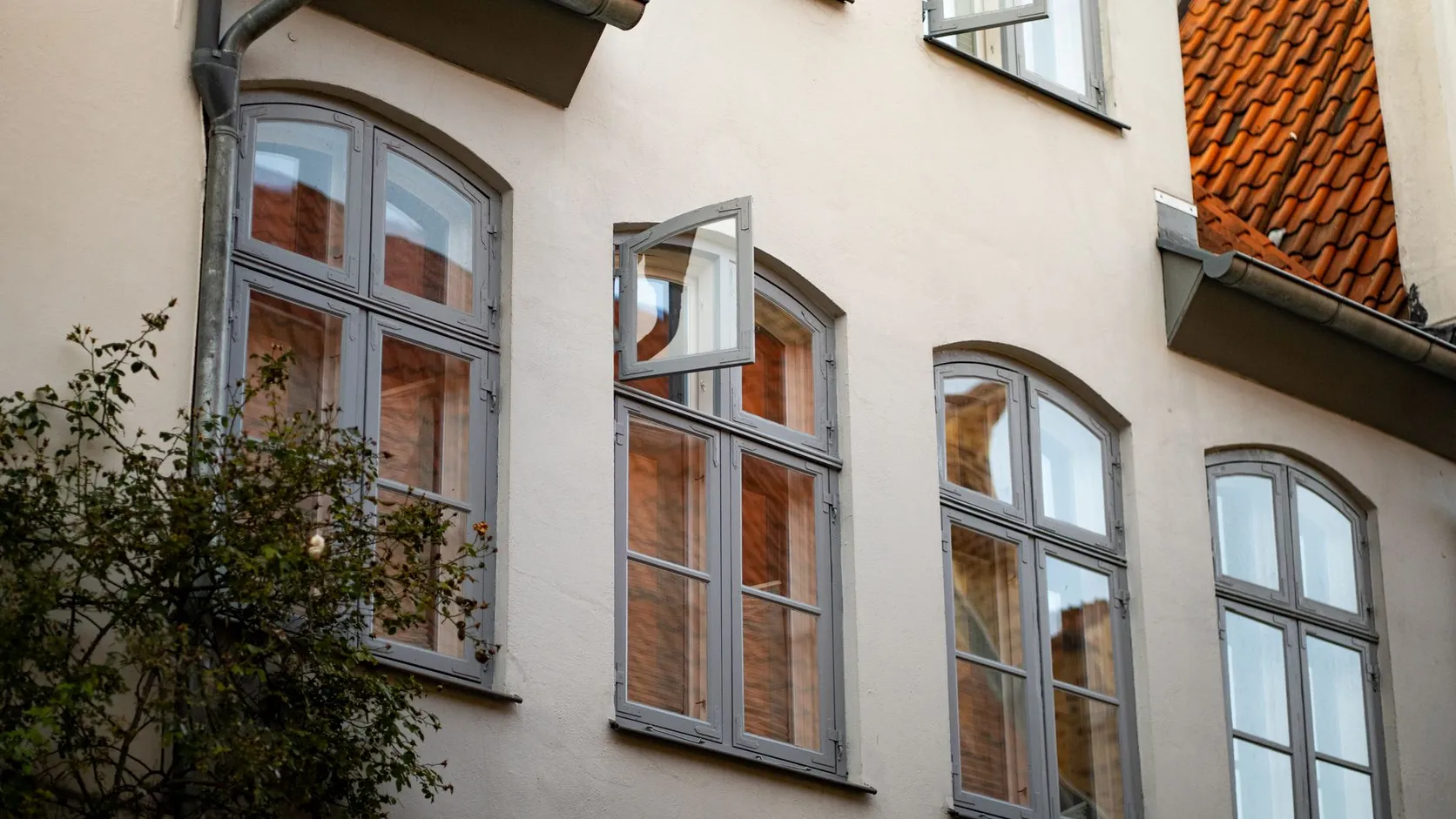 Façade claire aux fenêtres cintrées grises, dont un battant entrouvert, sous un toit de tuiles rouges