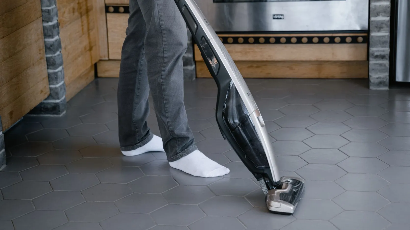 Personne en chaussettes blanches passant un aspirateur balai sur un carrelage hexagonal gris dans une cuisine