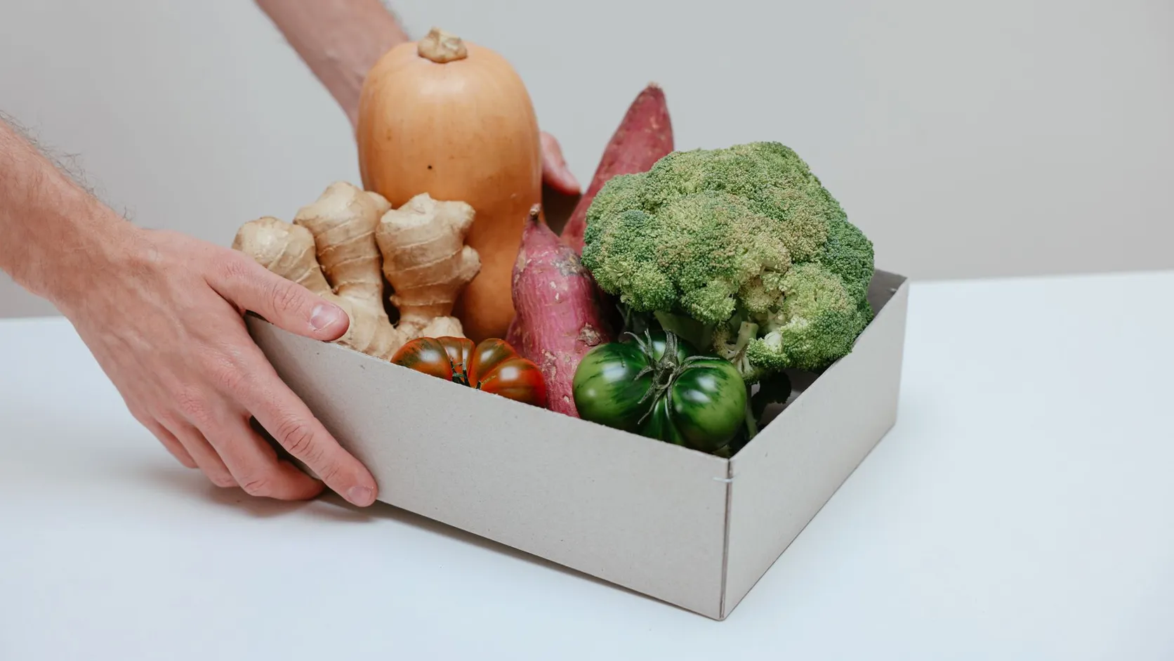 Mains posant une boîte en carton remplie de légumes : butternut, patates douces, gingembre, brocoli et tomates