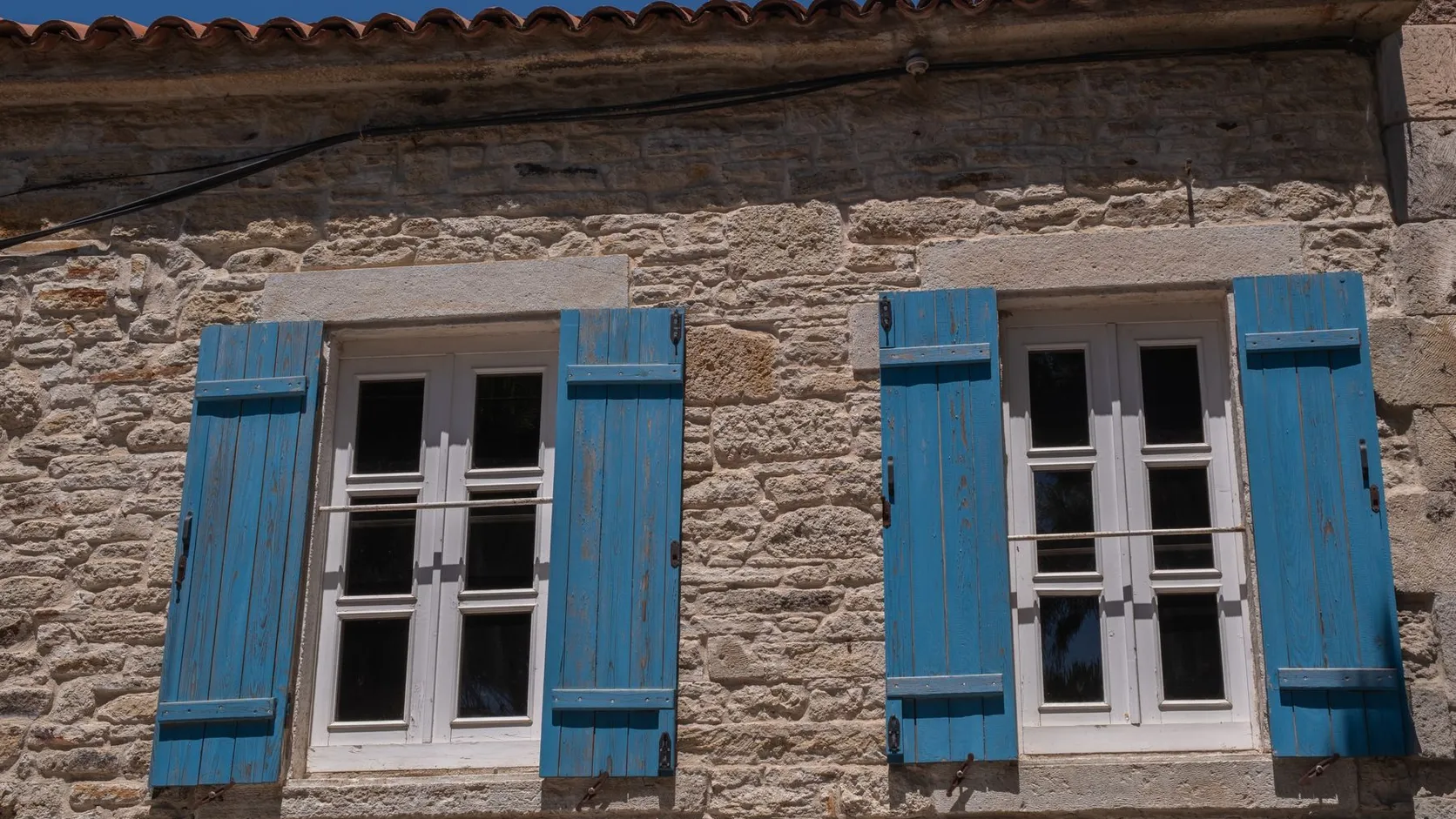 Façade en pierre avec deux fenêtres à volets bleus sous une avancée de toit en tuiles