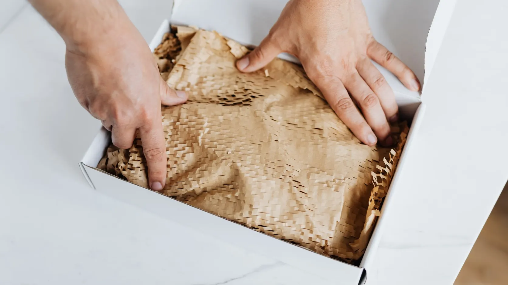 Deux mains glissant un papier kraft alvéolé de calage dans une boîte en carton blanche
