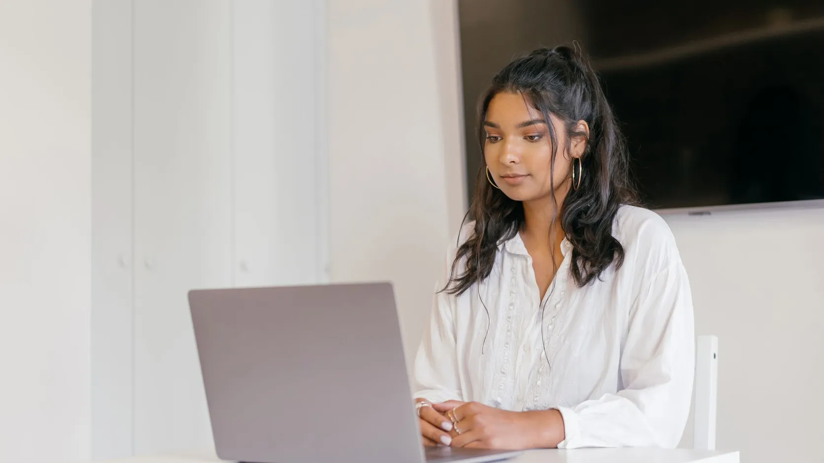 Jeune femme en chemisier blanc, concentrée devant un ordinateur portable posé sur une table blanche