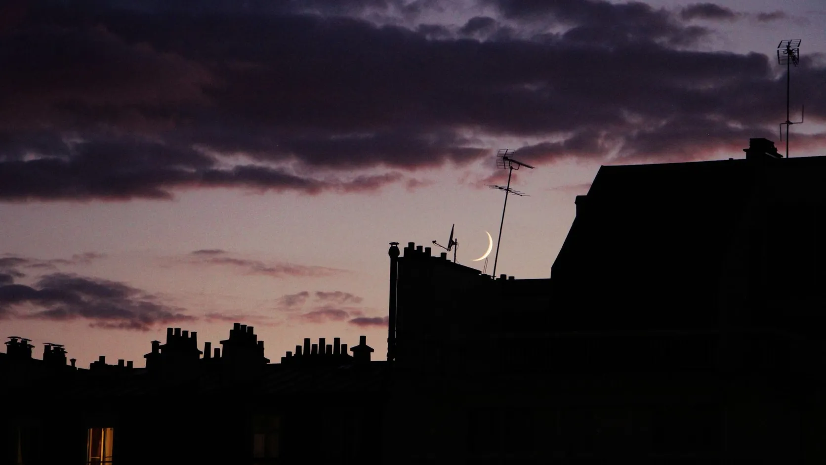 Toits d'immeubles en silhouette au crépuscule, surmontés de cheminées, d'une parabole et d'antennes râteau, avec un croissant de lune