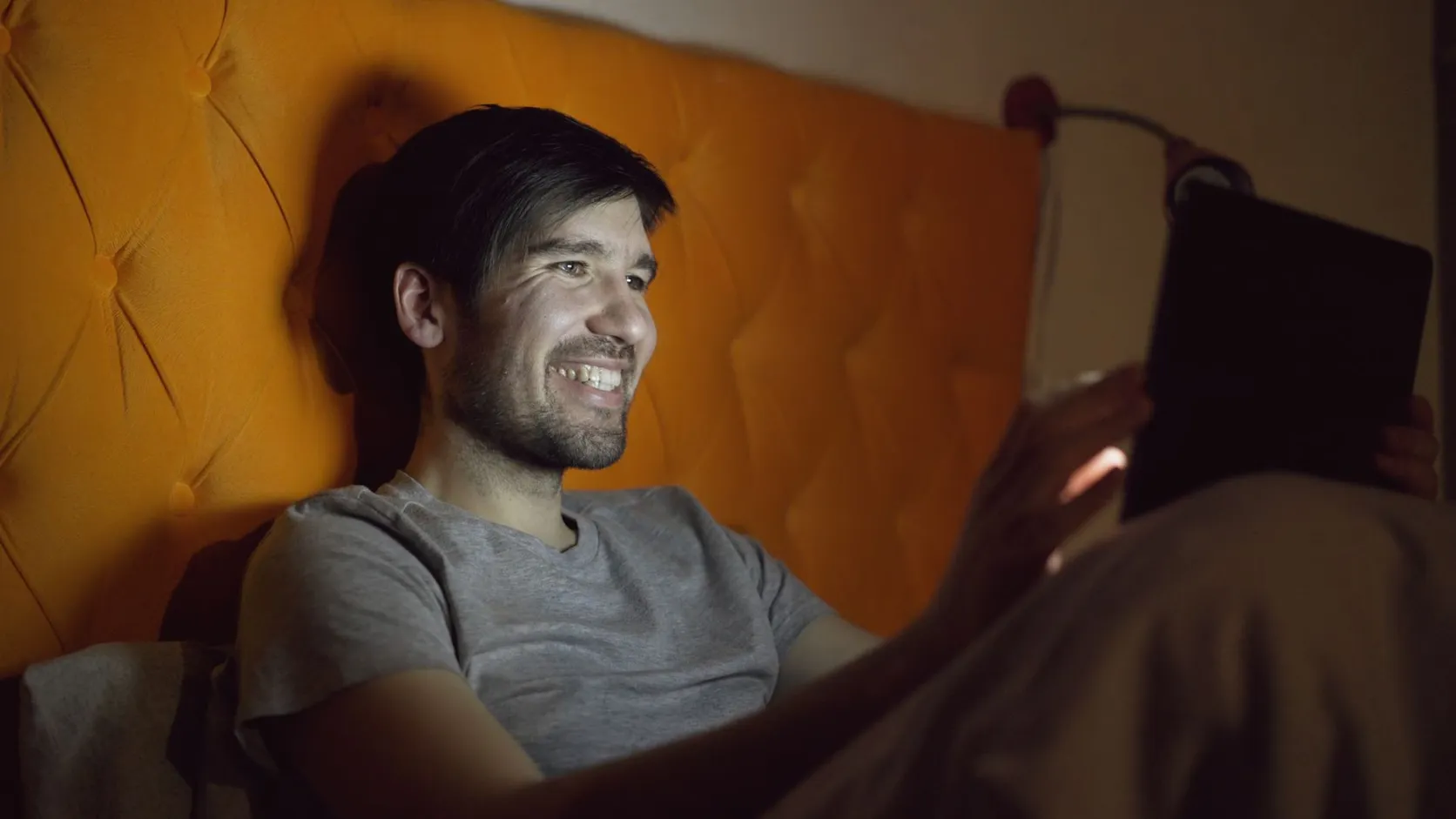 Homme souriant allongé dans son lit la nuit, le visage éclairé par l'écran de la tablette qu'il tient