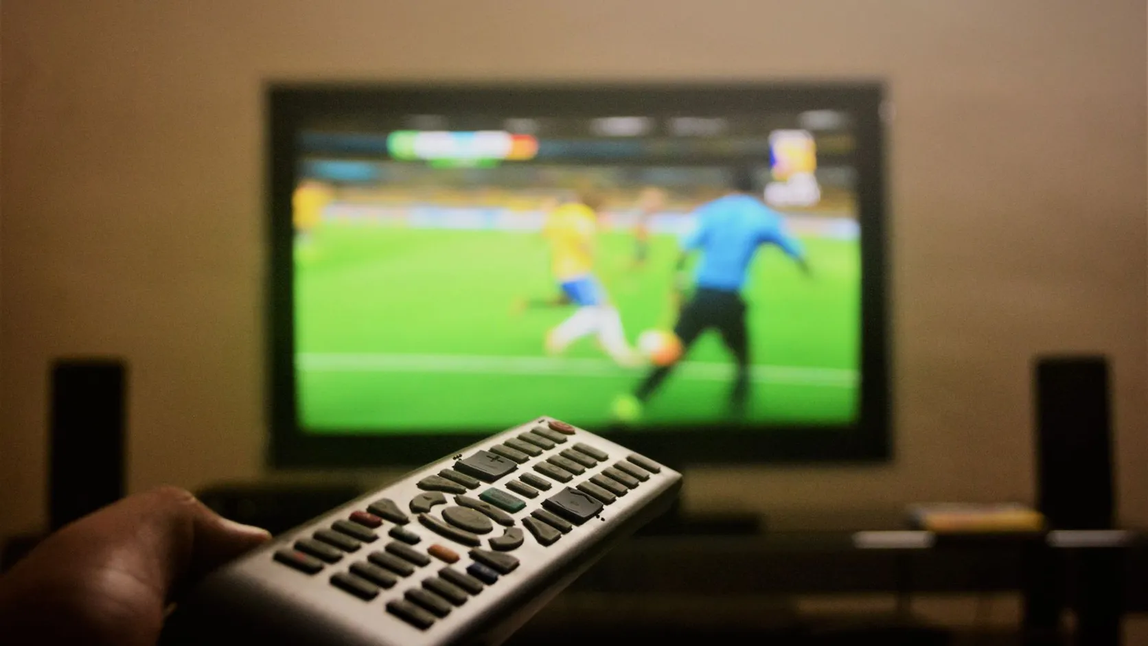 Main tenant une télécommande pointée vers un téléviseur flou qui diffuse un match de football
