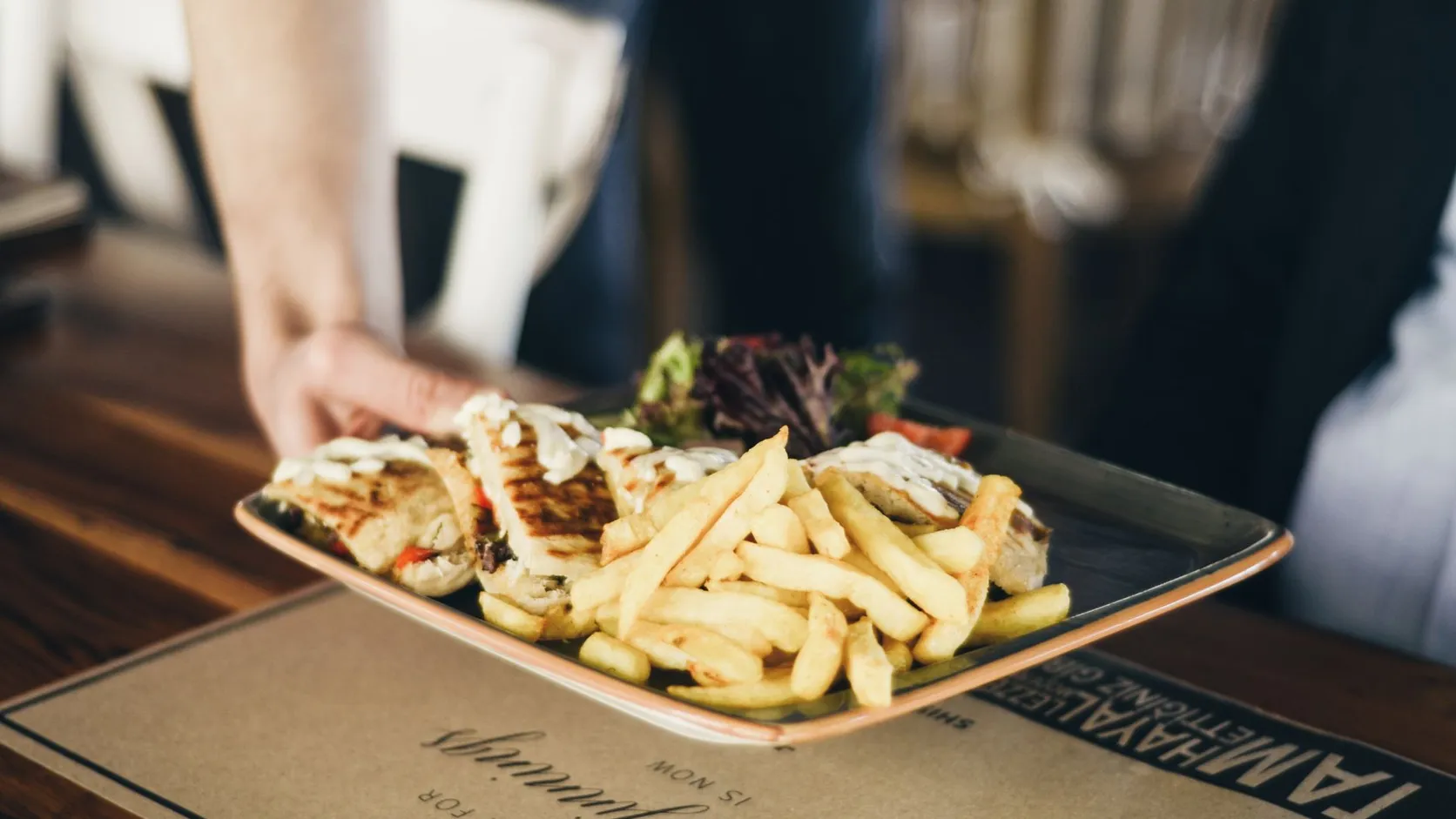 Plateau de sandwichs grillés, frites et salade tendu par une main au-dessus d'une table de restaurant en bois
